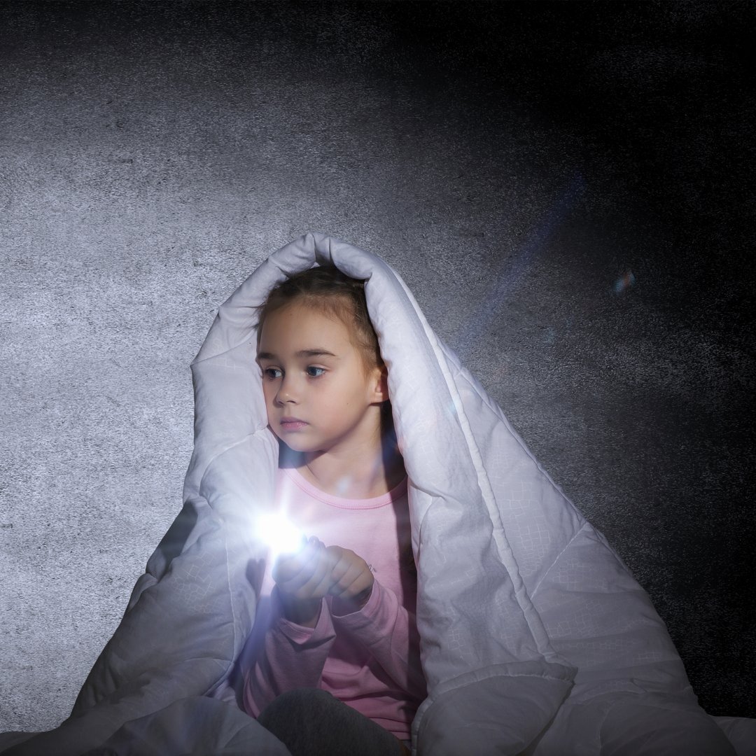 Imagem de uma menina embaixo de coberta, segurando uma lanterna