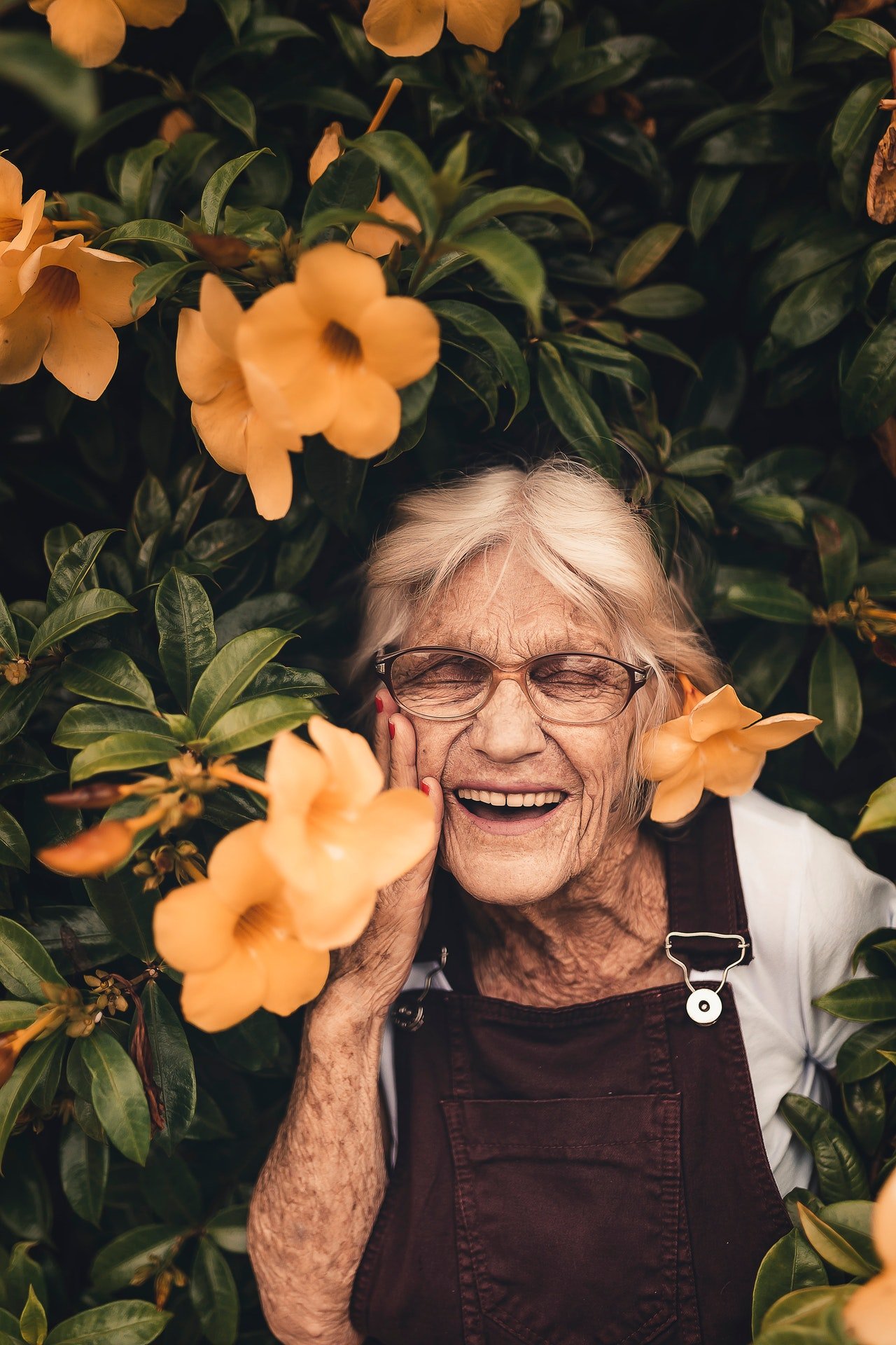 Senhora idosa em pé, rindo, em meio a um muro de flores.