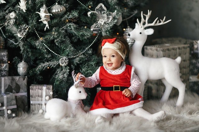 Bebê com roupa natalina em frente de caixas de presentes, decorações e árvore de natal.