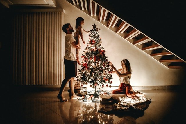 Família decorando árvore de Natal, com homem segurando menina para o alto para ela colocar uma estrela no topo da árvore.