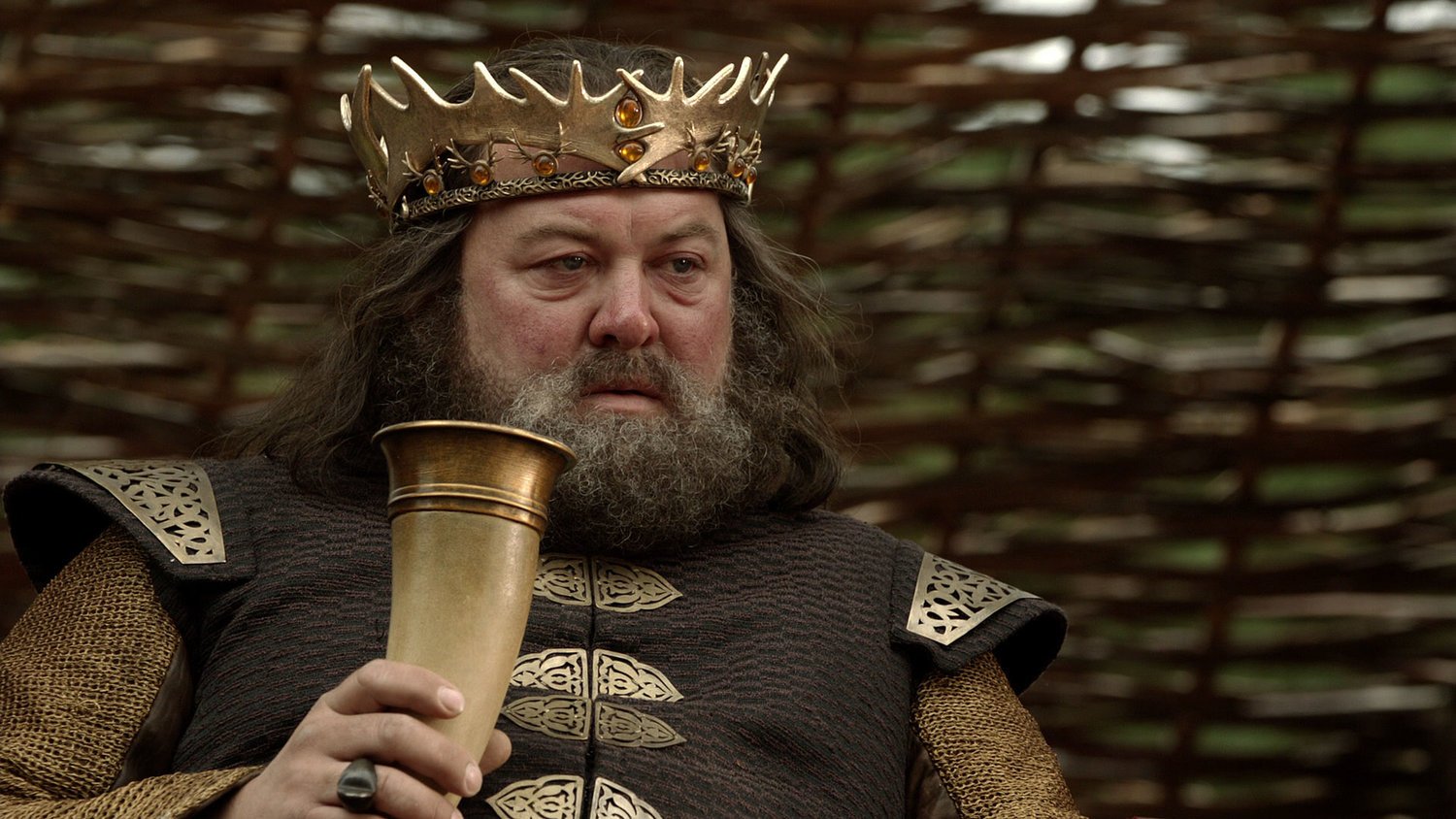 Robert Baratheon segurando uma taça na mão vestido com uma coroa na cabeça