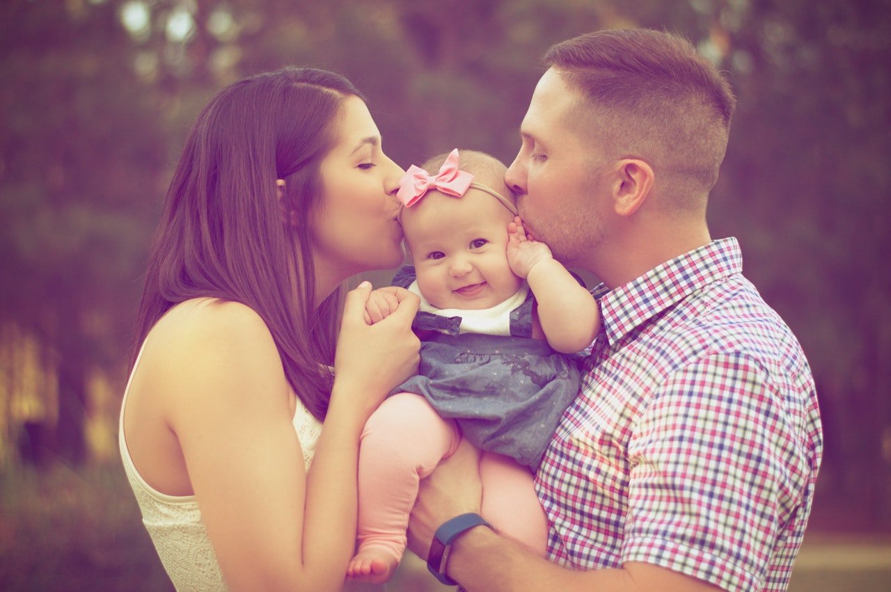 Casal segura uma bebê, e cada um beija um lado de seu rosto.