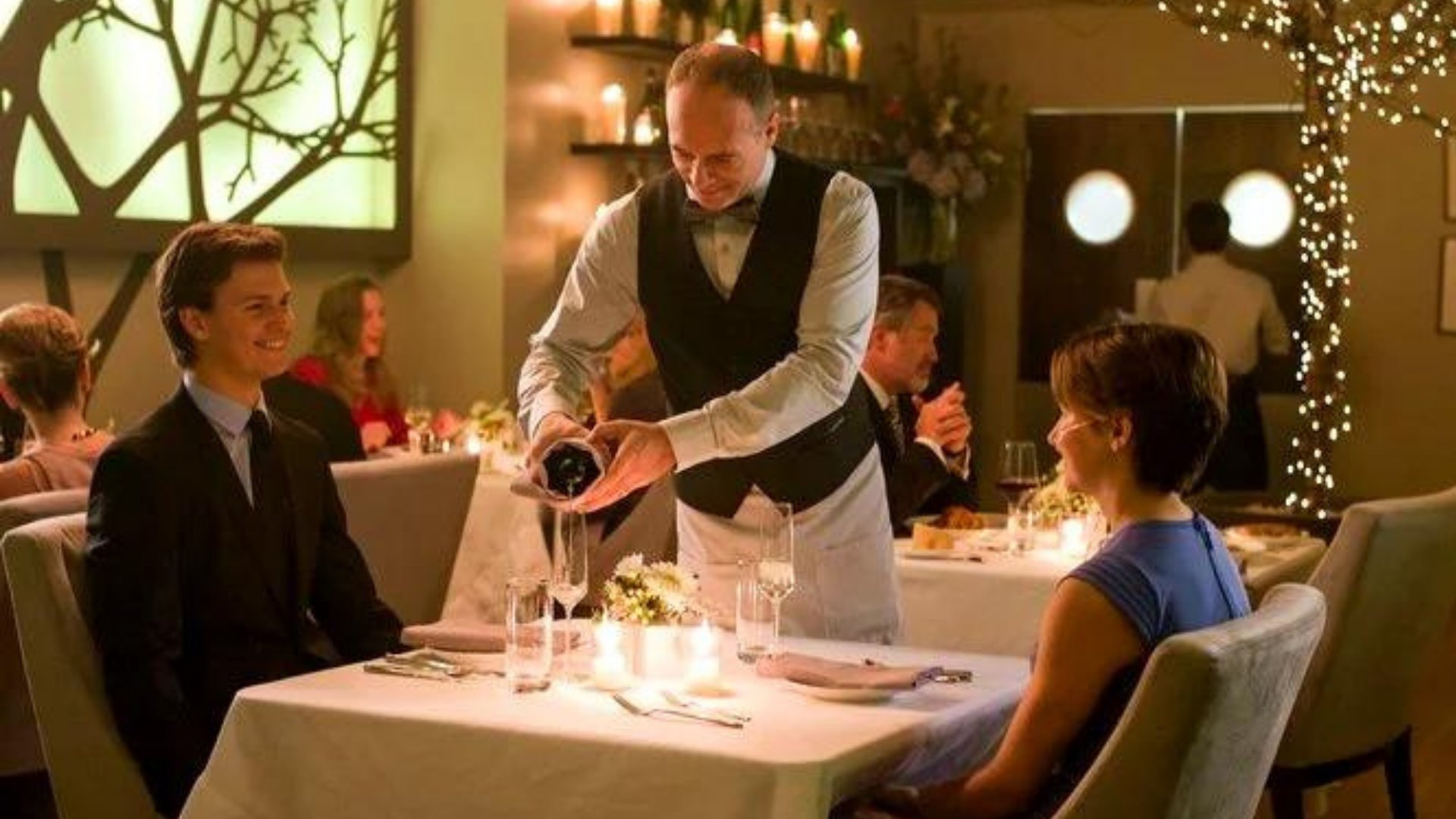 Gus e Hazel sentados na mesa de um restaurante enquanto um garçom serve champanhe para os dois