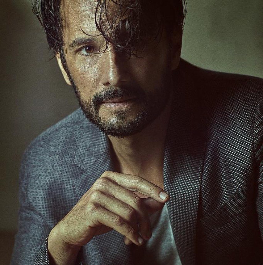 Rodrigo Santoro em sessão de fotos, com o cabelo levemente caído no rosto, e a mão solta em frente ao peito.