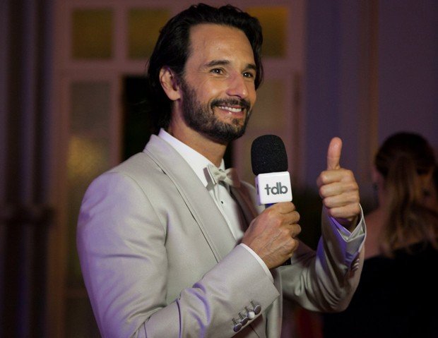 Rodrigo Santoro segurando um microfone com uma mão, e apontando o polegar da outra mão para cima, sorrindo.