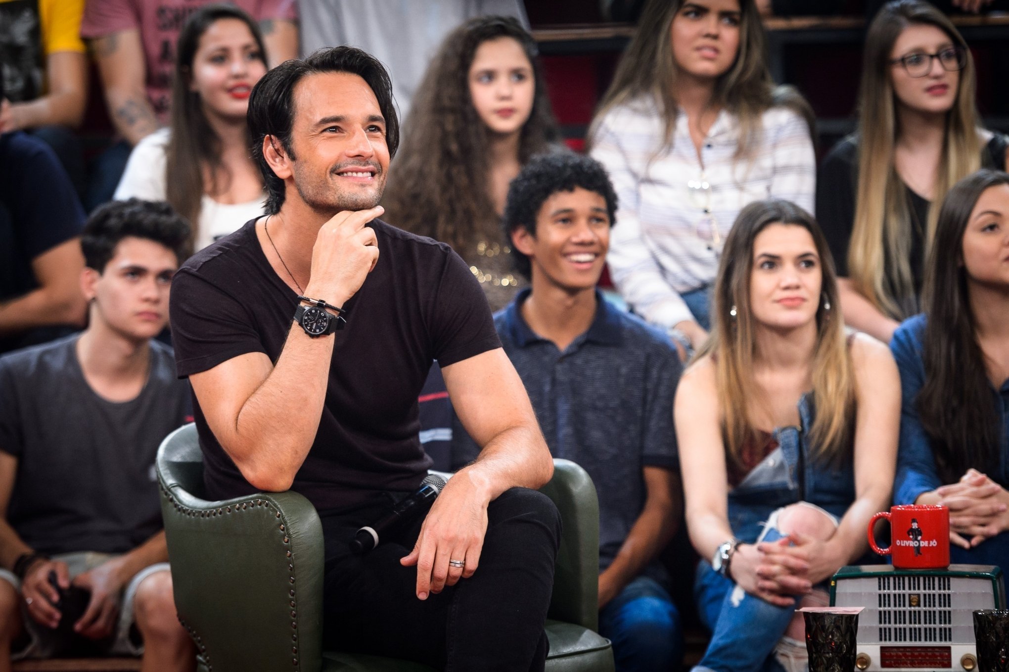 Rodrigo Santoro sorrindo e apoiando o rosto em uma das mãos durante programa de auditório.