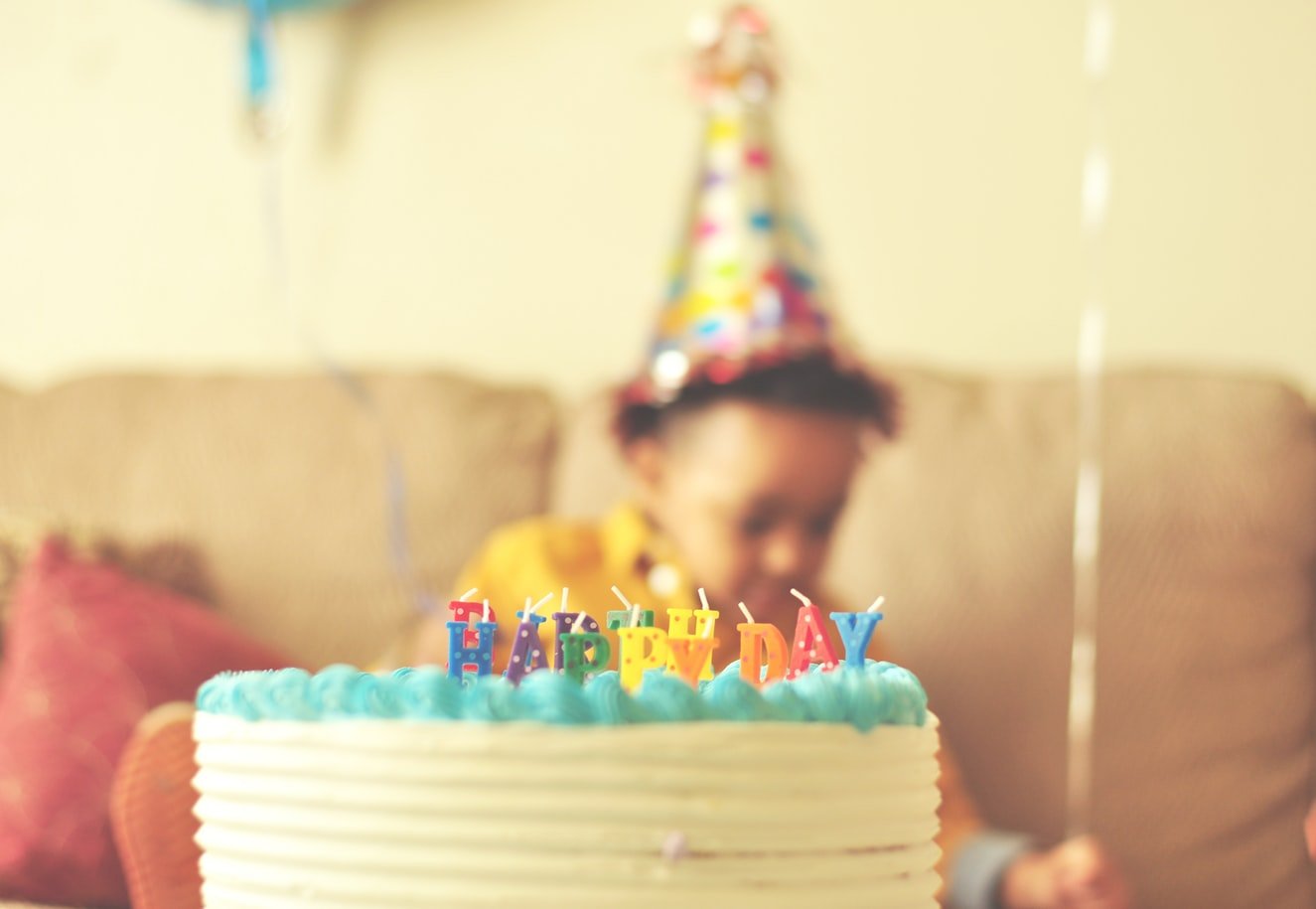 Criança com chapéu de festa segurando um balão atrás de um bolo de aniversário.