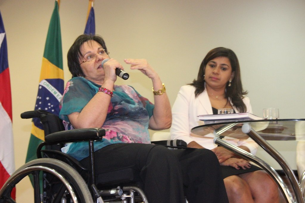 Maria da Penha segurando microfone e falando em sua cadeira de rodas ao lado de outra mulher