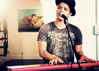 Cantor Bruno Mars vestindo camiseta e chapéu, tocando um teclado vermelho e cantando.