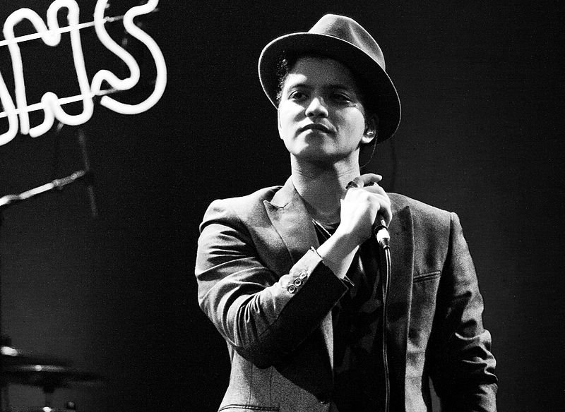 Bruno Mars vestindo terno, gravata e chapéu, segura um microfone durante um show.