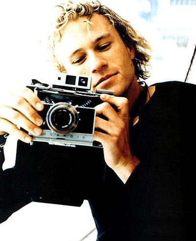 Imagem do ator Heath Ledger segurando uma câmera fotográfica.