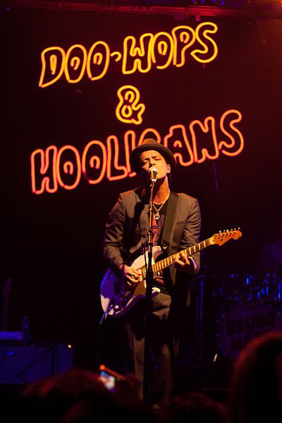 Bruno Mars se apresentando em show, cantando e tocando guitarra.