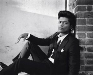 Divulgação - Vagalume Bruno Mars sentado no chão, encostado em um muro, com os joelhos flexionados e o braço direito apoiando no joelho direito.