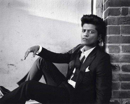 Bruno Mars sentado no chão, encostado em um muro, com os joelhos flexionados e o braço direito apoiando no joelho direito.