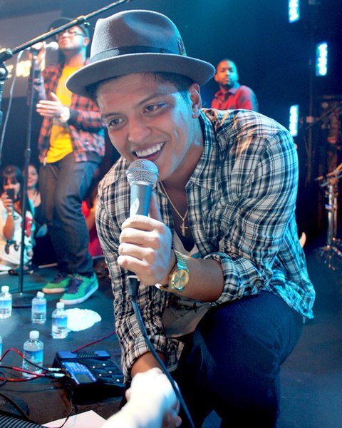 Cantor Bruno Mars em show, ajoelhado na beirada do palco, segurando a mão de uma fã enquanto canta.