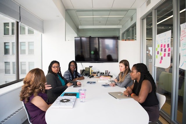 Mulheres fisicamente diferentes sentadas ao redor de uma mesa de reunião, conversando.