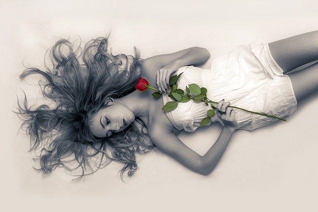 Mulher deitada, usando um macacão curto, com seu cabelo espalhado para todos os lados. A foto é em preto e branco, e ela segura uma rosa com a mão direita, sobre seu peito. A rosa é o único elemento na foto que está colorido.