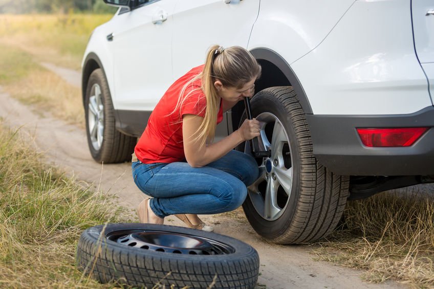 Mulher trocando pneu traseiro de carro em estrada de terra.