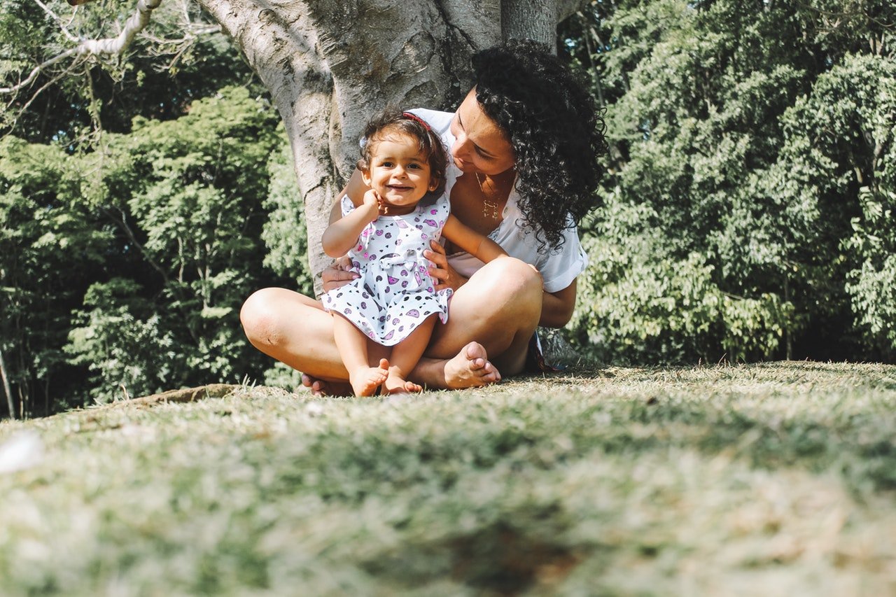 Mulher sentada embaixo de árvore com bebê em seu colo. A bebê sorri com uma mão no rosto, enquanto a mulher olha para ela.