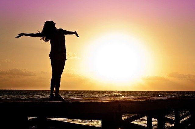 Silhueta de mulher de pé em um pier, de frente pra o mar e o sol, com os braços abertos e a cabeça inclinada para trás.