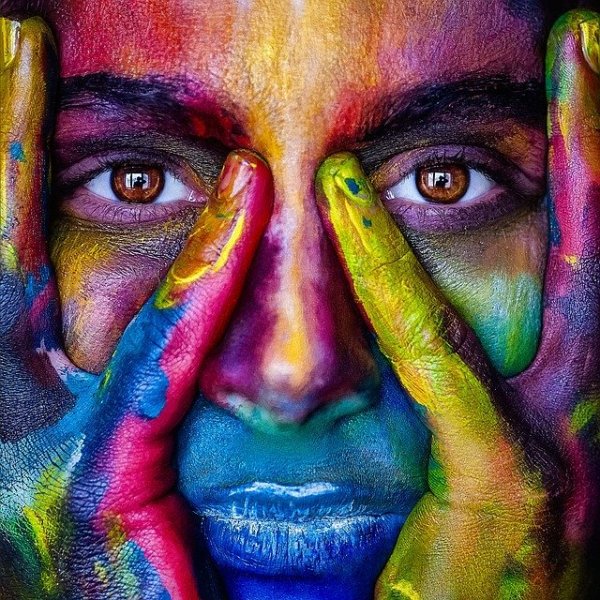 Pixabay/Alexandr Ivanov Mulher com rosto e mãos pintadas de múltiplas cores em tons de degradê. Ela olha para a câmera e tem seus mindinhos apoiados em seu nariz, enquanto os dedos anelares estão na lateral do rosto.