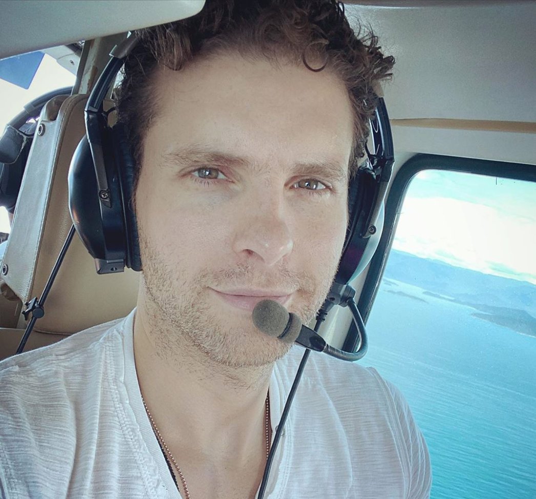 Thiago Fragoso dentro de helicóptero, usando fone de ouvido com microfone, sobrevoando o mar.