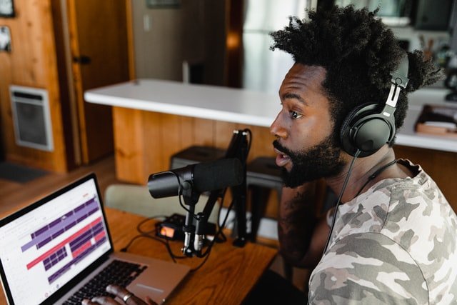 Homem negro usando fones de ouvido, em frente à microfone e computador.