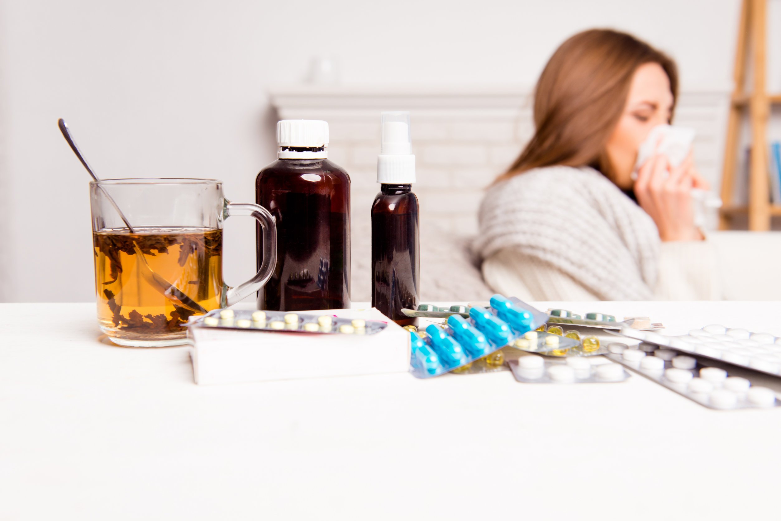 Vários remédios e xaropes sobre uma mesa com mulher gripada assoando o nariz no fundo