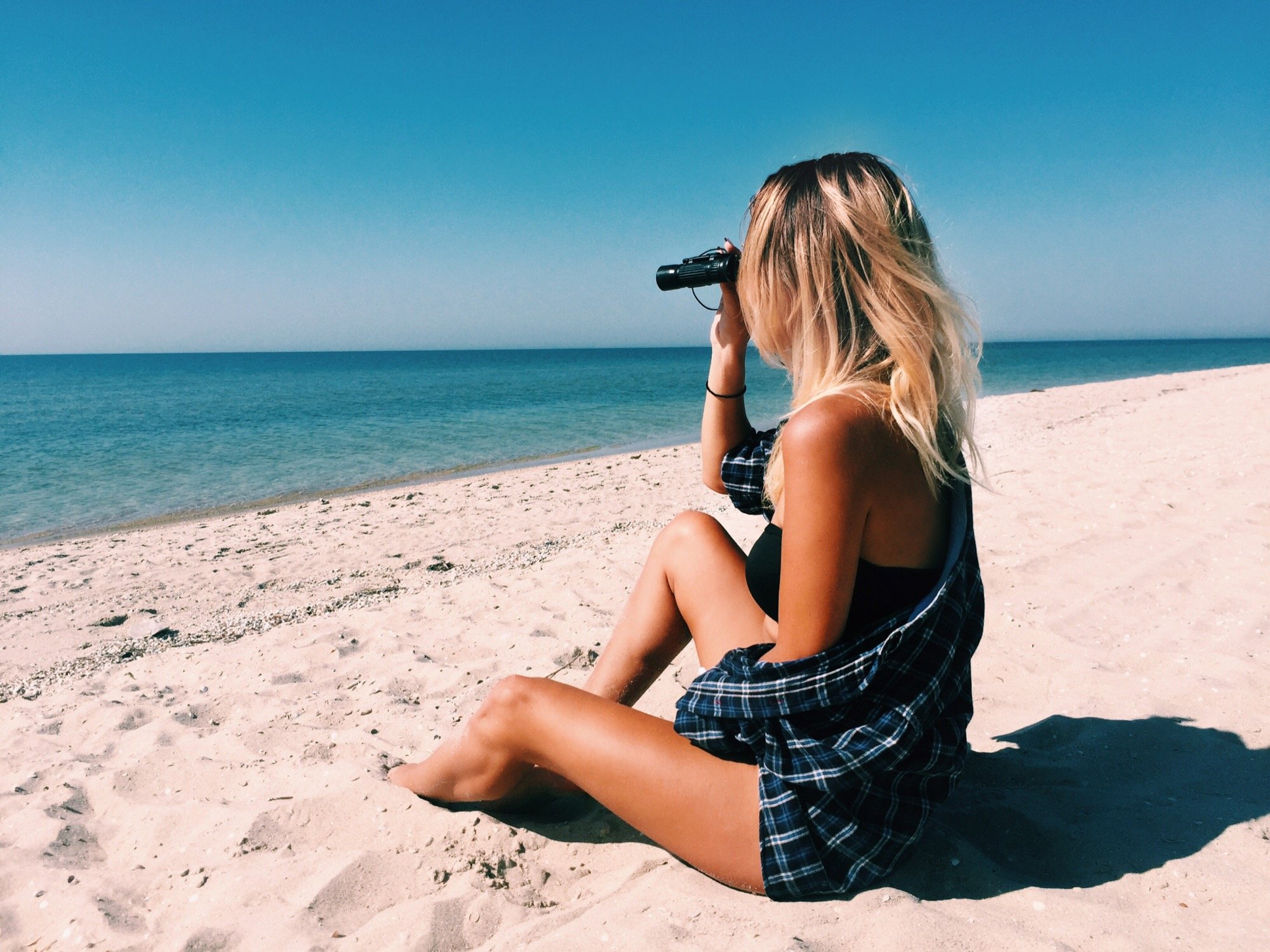 Mulher sentada na areia da praia observando com binóculo