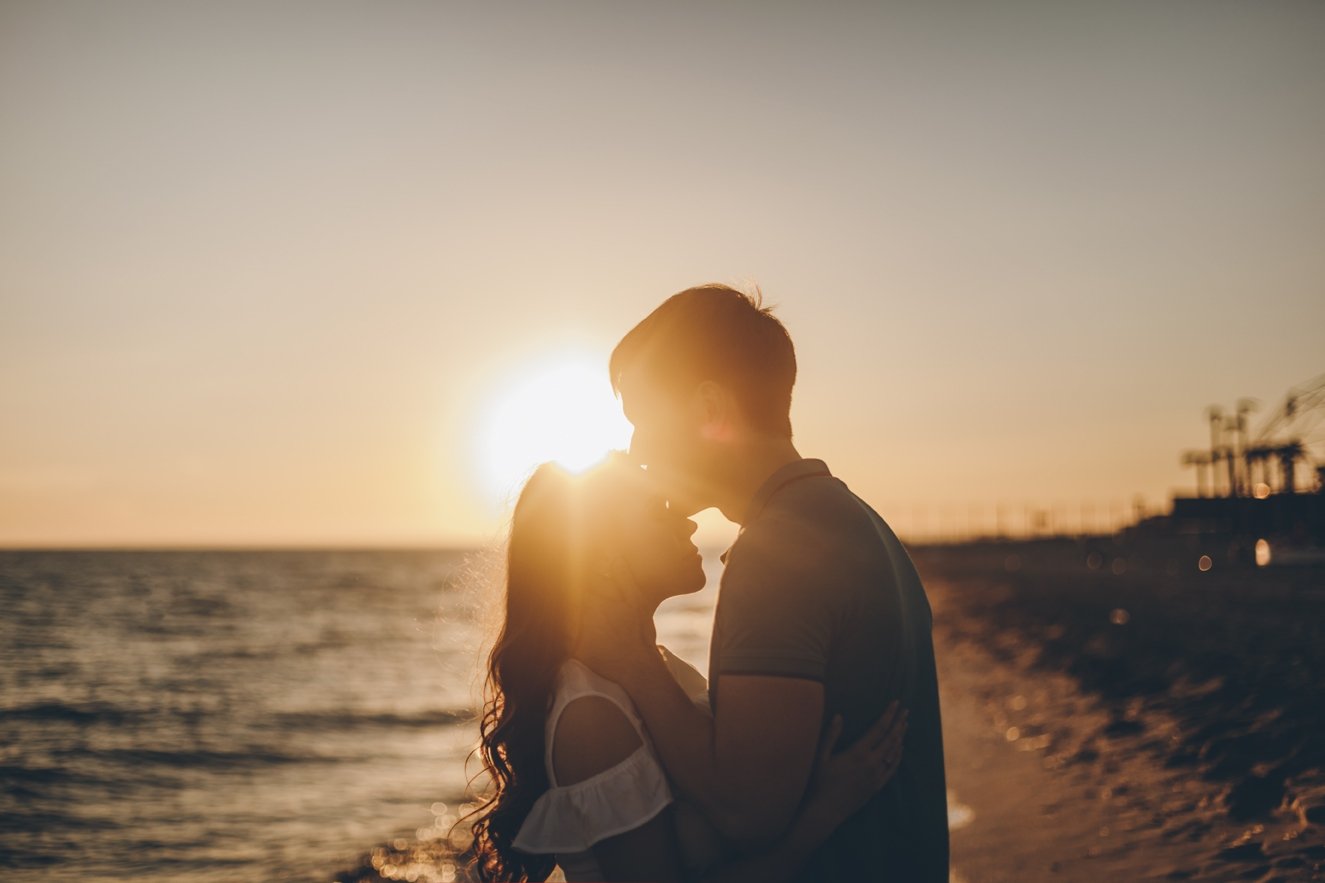 Homem beijando testa de mulher a sua frente ao lado de uma praia e o pôr do sol