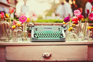 Maquina de escrever verde em cima de móvel antigo, rodeada por copos com flores