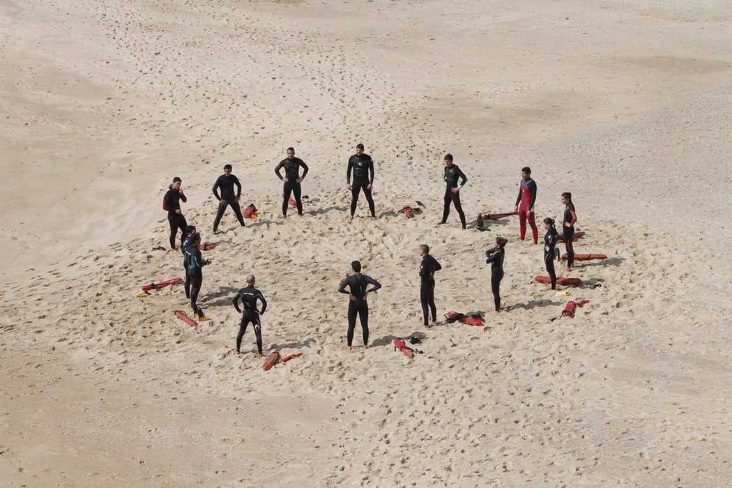 Pessoas em pé na praia, vestindo roupa de neoprene, formam um circulo.