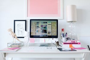 Escritório branco e rosa, com computador, luminária de chão, canetas e cadernos