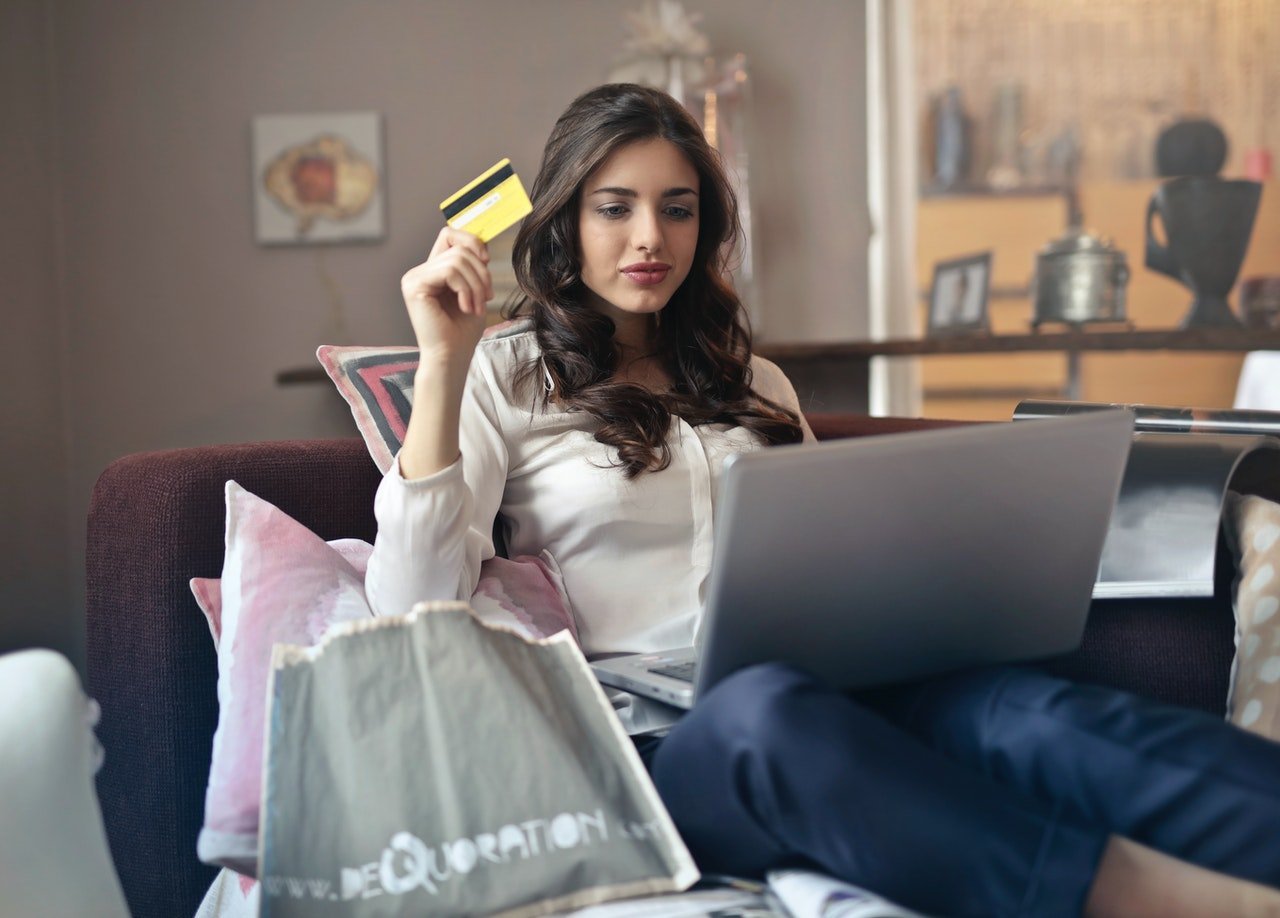 Mulher branca sentada com notebook no colo e sacolas de compras ao lado.