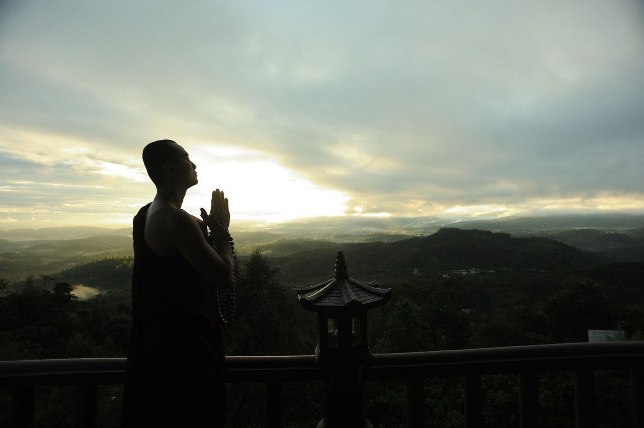 Monge rezando em um templo no topo de uma montanha, com vista para montanhas menores e uma pequena cidade.