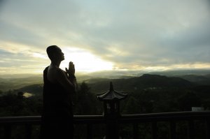 Pexels - nhan thai Monge rezando em um templo no topo de uma montanha, com vista para montanhas menores e uma pequena cidade.