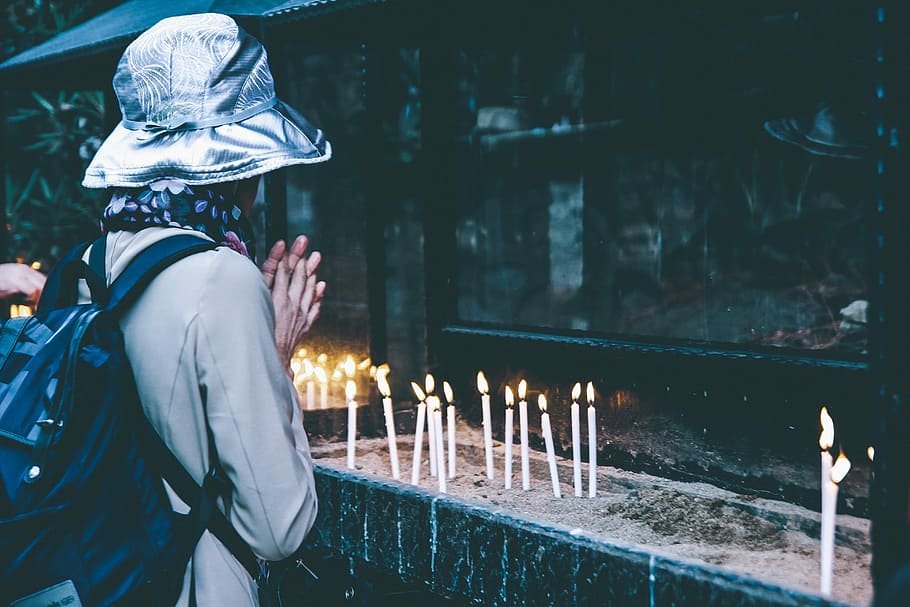 Pessoa de costas para a câmera, usando um chapéu, tem suas mãos unidas em frente do seu corpo. A sua frente, muitas velas acesas.