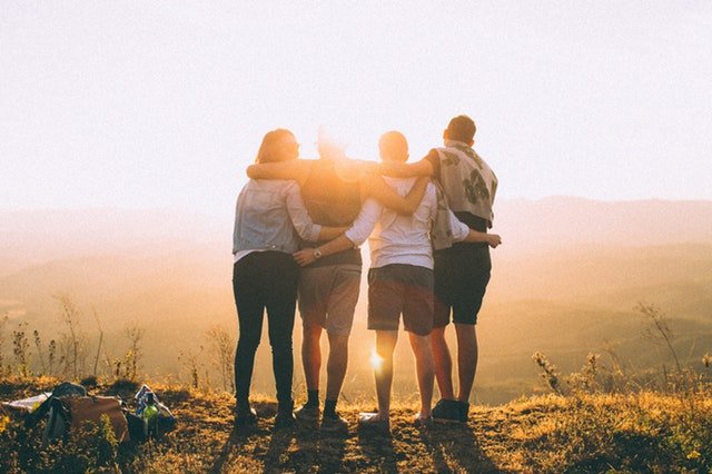 Quatro amigos abraçados lado a lado em frente ao pôr do sol.