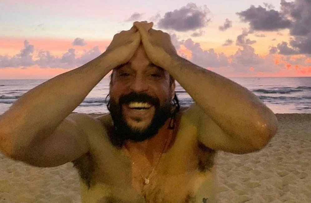 Homem branco de barba preta com as mãos na cabeça, e expressão sorridente.