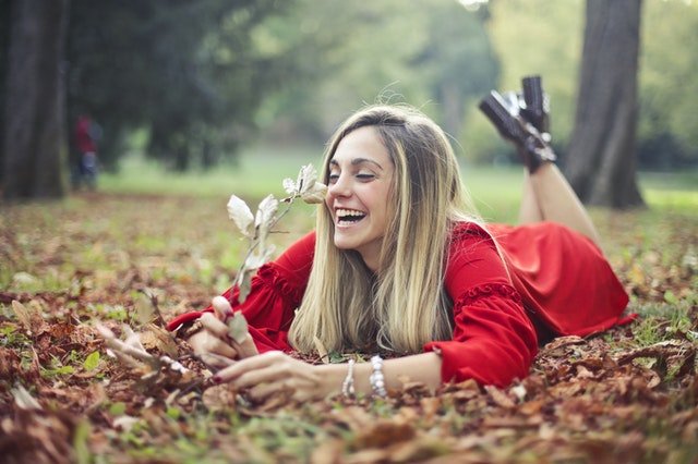 Mulher deitada em folhas secas, com a barriga para baixo, segurando uma flor branca e rindo.