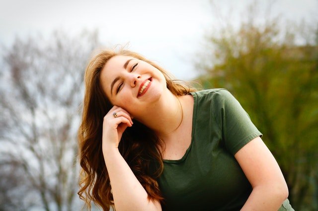 Mulher sorrindo com a mão direita em sua bochecha, em um parque.