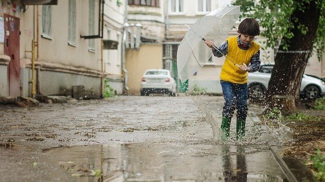 Menino segurando guarda-chuva transparente, enquanto pula em uma poça de água.