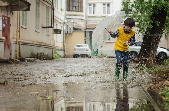 Menino segurando guarda-chuva transparente, enquanto pula em uma poça de água.