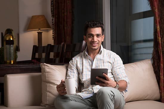 Homem sentado no sofá sorrindo olhando para frente, segurando tablet e xícara de café