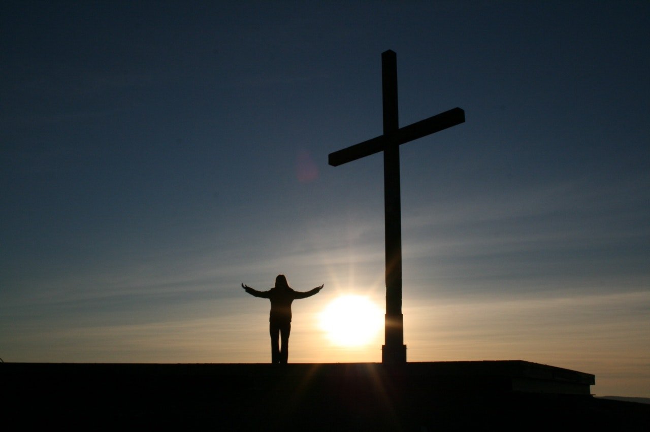 Silhueta de pessoa com os braços abertos, ao lado de uma cruz com o triplo de sua altura. Ao fundo, um céu escuro com o sol se pondo.