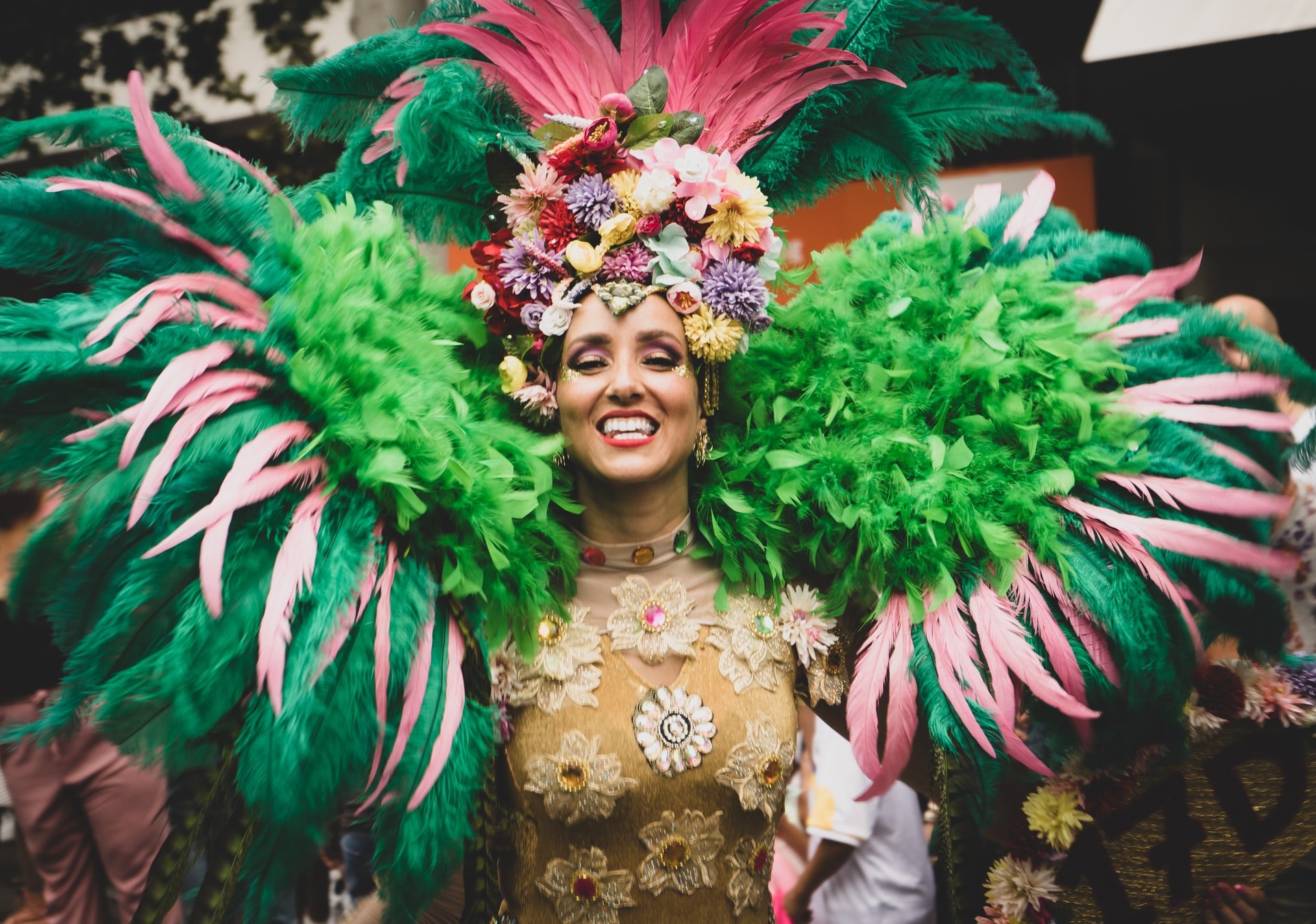 Mulher com fantasia carnavalesca grande, com ombreiras cheia de penas e chapéu grande, sorrindo para a câmera.