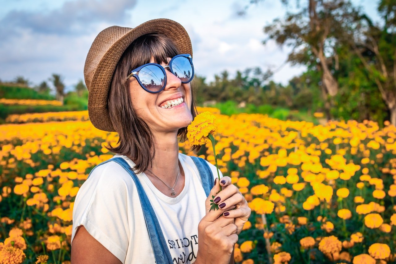 Mulher com chapéu e óculos de sol, em um campo de flores amarelas, sorrindo.