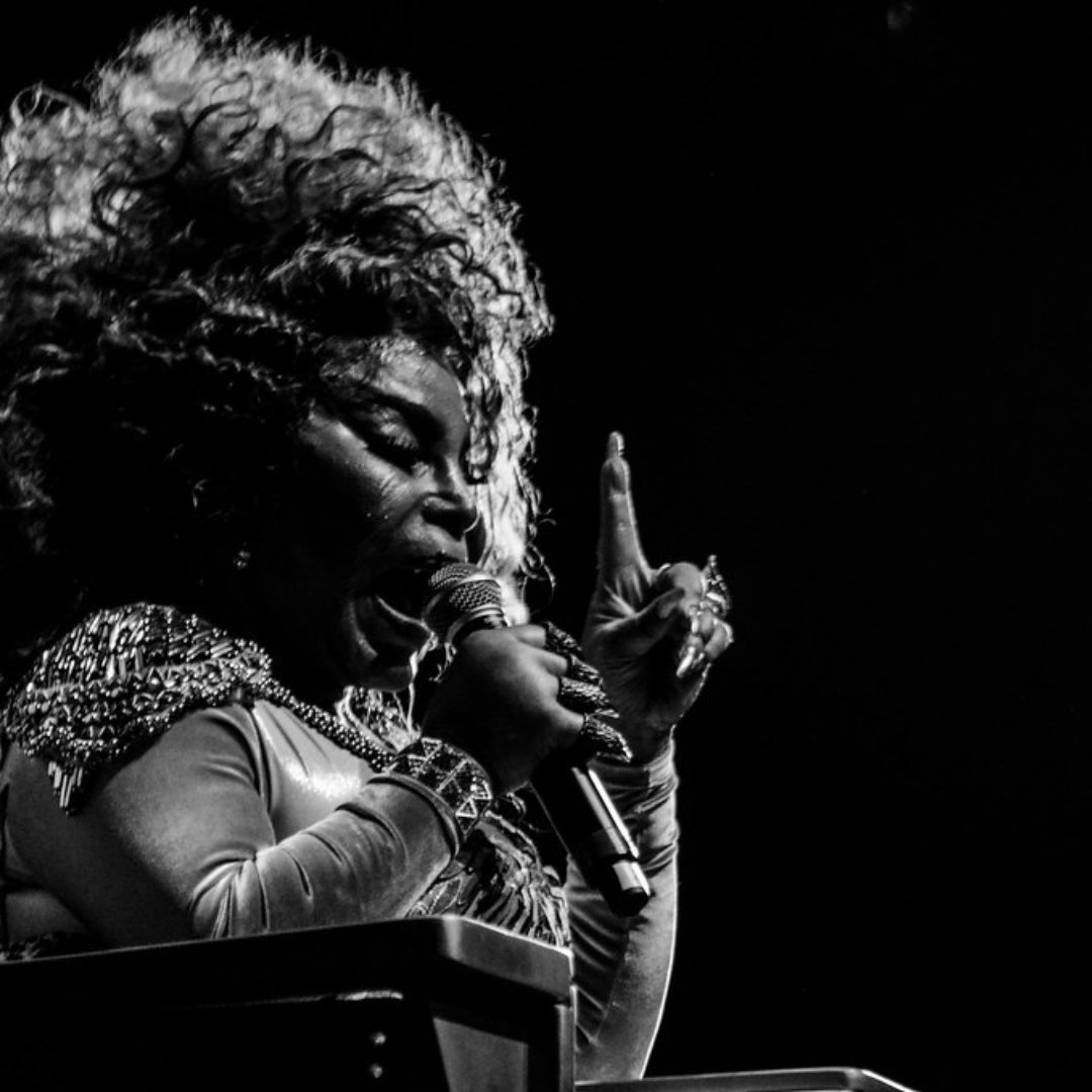 Imagem em preto e branco da cantora Elza Soares, que está cantando e apontando o dedo para cima