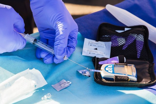 Pessoa com luvas azuis manuseando seringa e utensílios médicos.