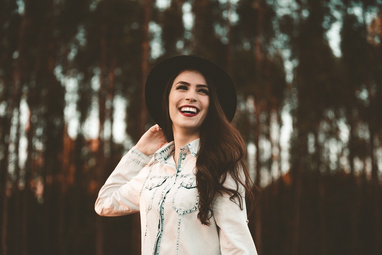 Mulher sorrindo usando chapéu preto e jaqueta jeans. Ao fundo, árvores altas e bem próximas.
